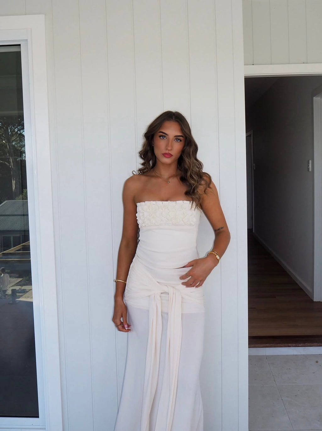 A woman modeling a strapless white maxi dress with a textured bodice and a long draped tie skirt, standing in front of a white paneled wall.