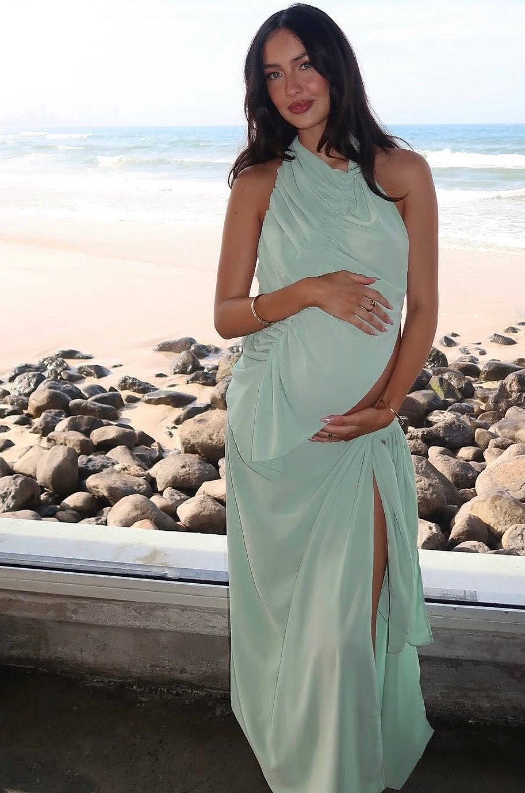 A pregnant woman wearing a light sage green halter-neck maxi dress stands on a rocky beach with the ocean in the background. - @ReinaTaia