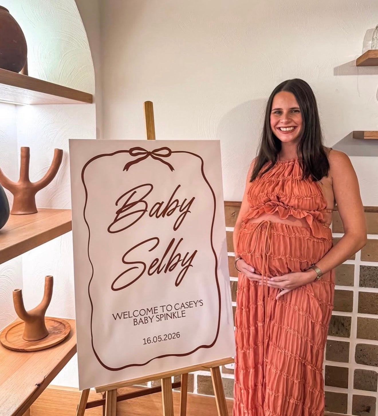 A smiling pregnant woman stands next to a custom baby shower sign on an easel in a rustic indoor setting. The sign reads 'Baby Selby' and 'WELCOME TO CASEYS BABY SPINNILE 16. - Caseyselby