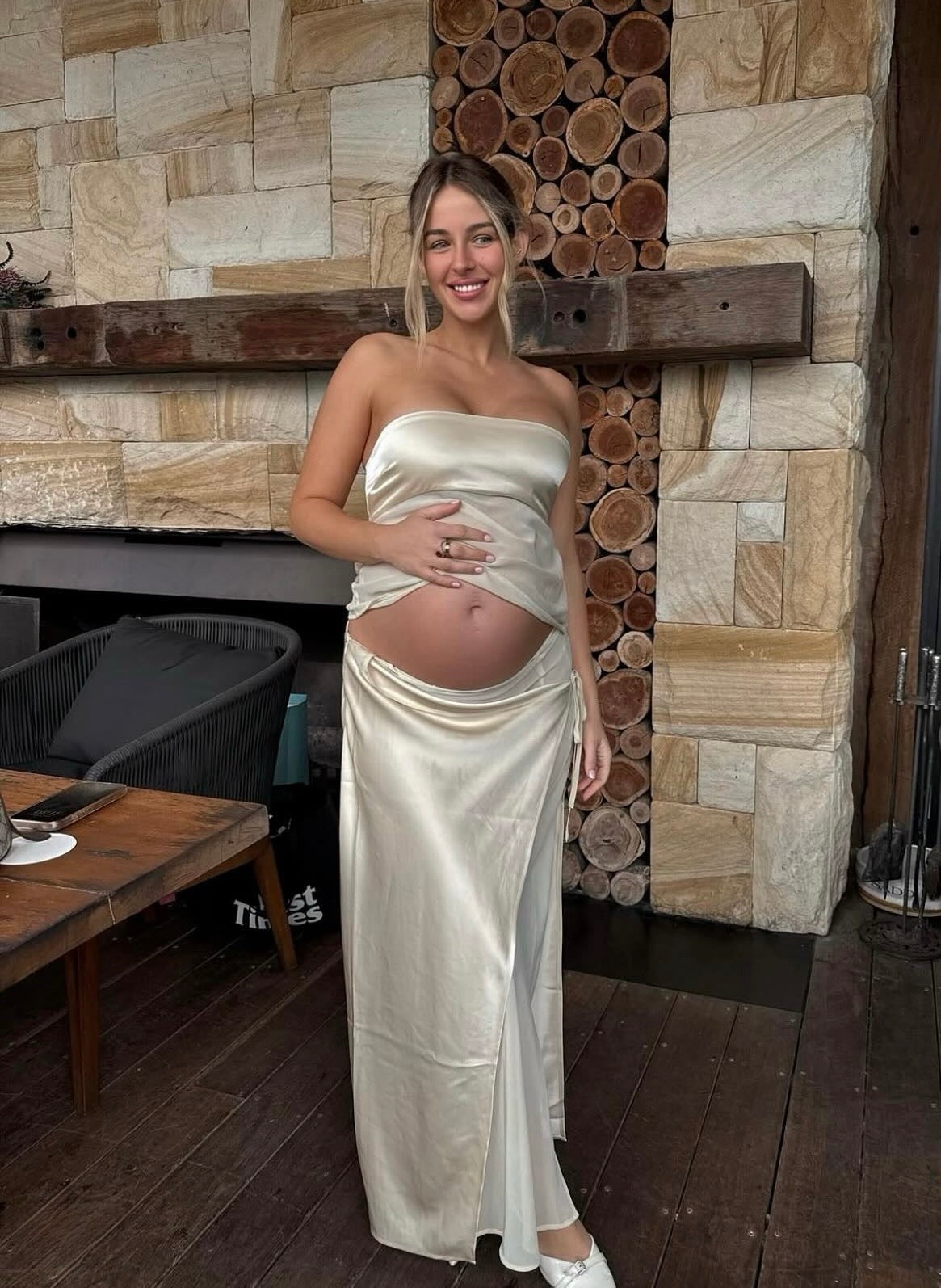 A pregnant woman wearing a strapless champagne satin maxi dress, standing in front of a rustic stone fireplace. - @georgiajanedavies