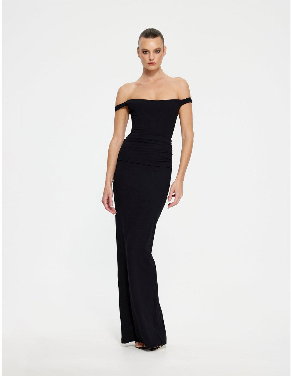 A woman modeling a black off-the-shoulder maxi dress with a fitted bodice and flared skirt, photographed against a plain light gray studio background.