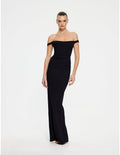 A woman modeling a black off-the-shoulder maxi dress with a fitted bodice and flared skirt, photographed against a plain light gray studio background.