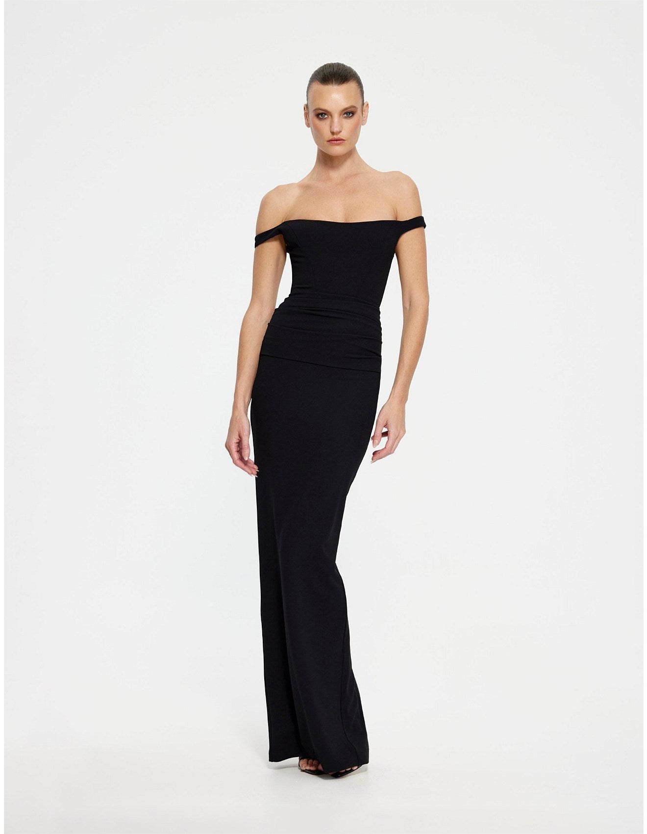 A woman modeling a black off-the-shoulder maxi dress with a fitted bodice and flared skirt, photographed against a plain light gray studio background.