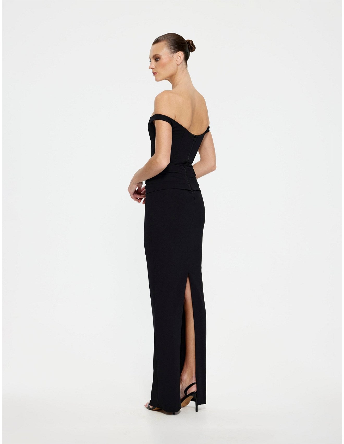 A woman modeling a black off-the-shoulder floor-length gown with a high front slit, shown from the back against a plain white studio background.