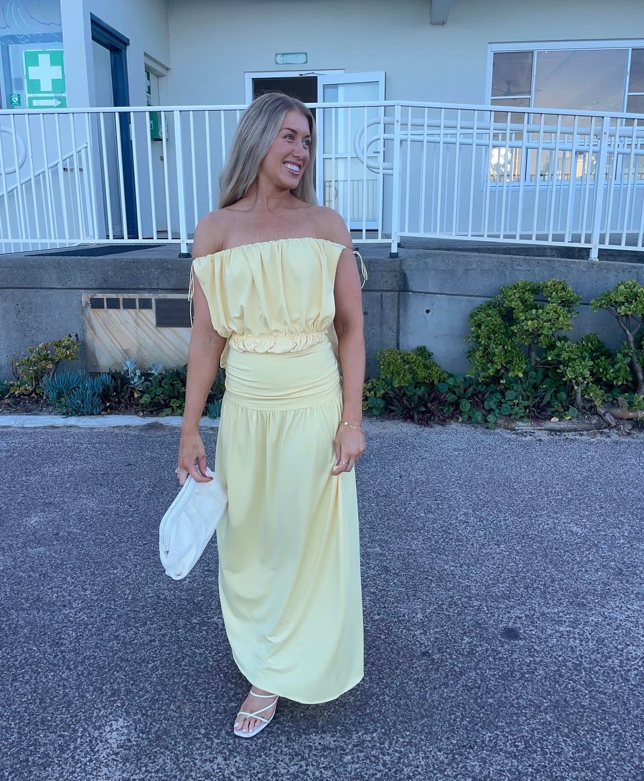 A woman wearing a pale yellow off-the-shoulder maxi dress with a ruffled waist, holding a white clutch and standing on a paved surface in front of a building with a white railing.