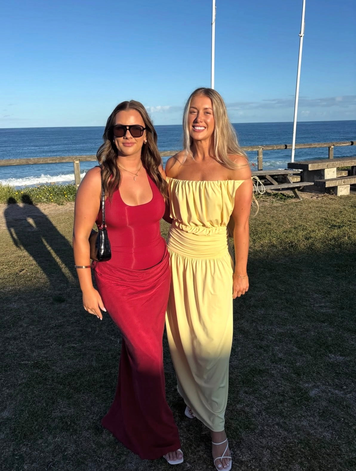 Two women posing outdoors by the ocean, wearing long sleeveless maxi dresses in red and pale yellow. One woman is wearing sunglasses, and the background shows the blue ocean, grassy ground, and a wooden fence.