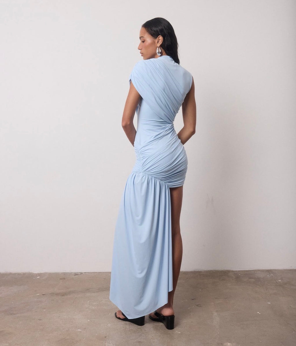 A woman stands with her back to the camera wearing a light blue draped maxi dress with a high slit and black heeled sandals against a plain off-white wall and concrete floor.