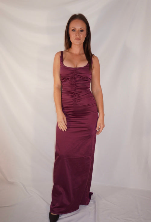 A woman modeling a sleeveless, floor-length ruched gown in a deep plum or burgundy shade, standing against a wrinkled white studio backdrop.