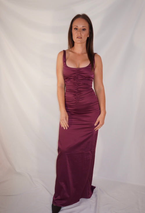 A woman modeling a sleeveless, floor-length ruched maxi dress in a deep plum purple color, standing against a white fabric backdrop.