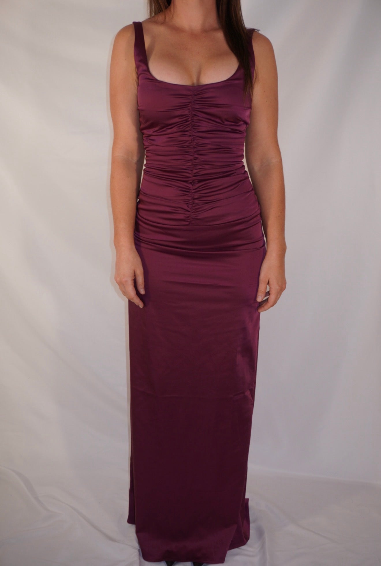 A woman wearing a sleeveless, floor-length ruched maxi dress in a deep plum or burgundy shade, standing against a white fabric backdrop.