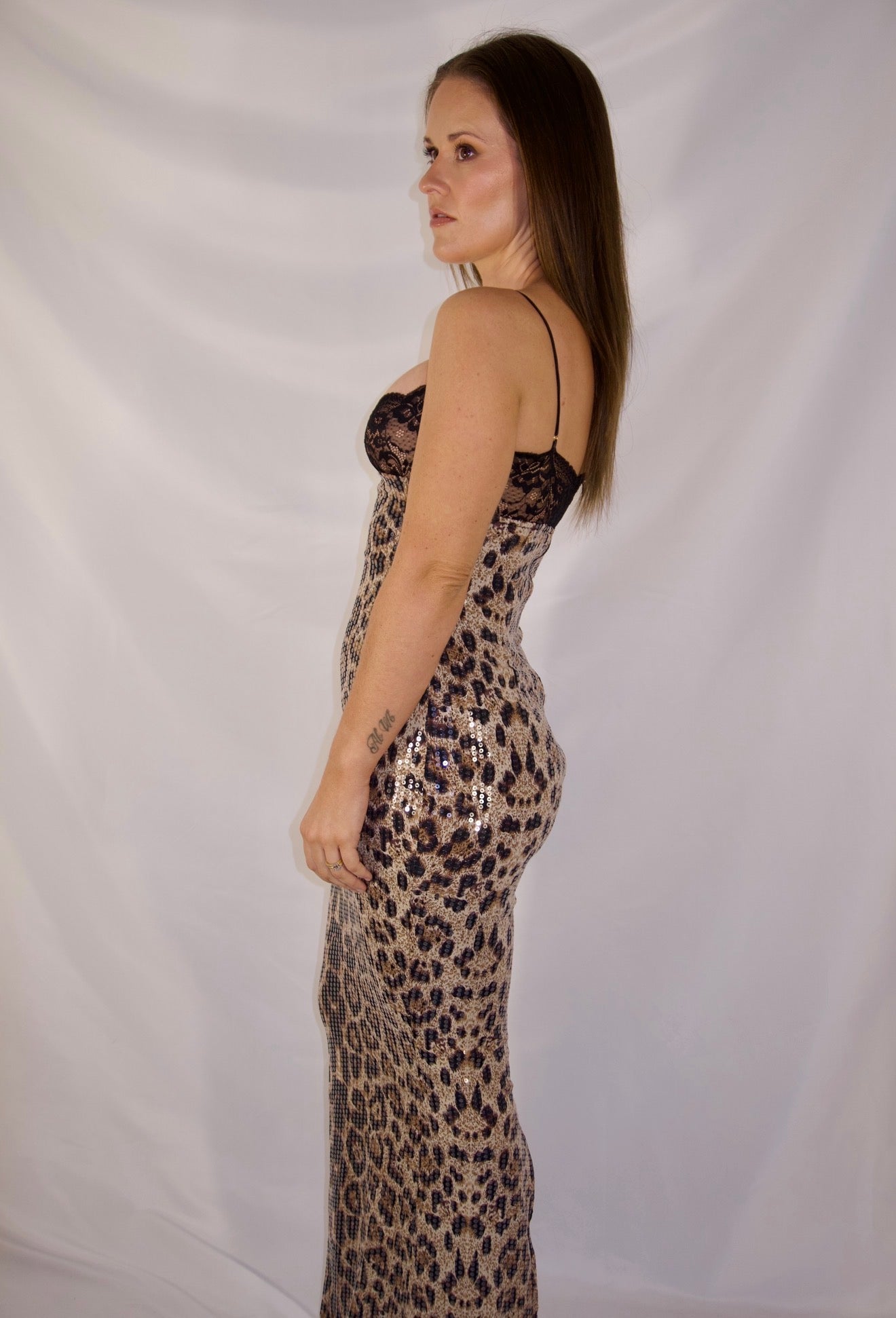 A woman modeling a floor-length leopard print dress with thin straps, standing in profile against a white draped fabric backdrop.