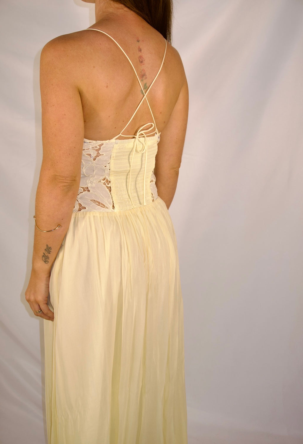 A woman shown from the back wearing a pale yellow maxi dress with a lace corset-style back bodice and thin tie-back straps, photographed against a plain light backdrop.
