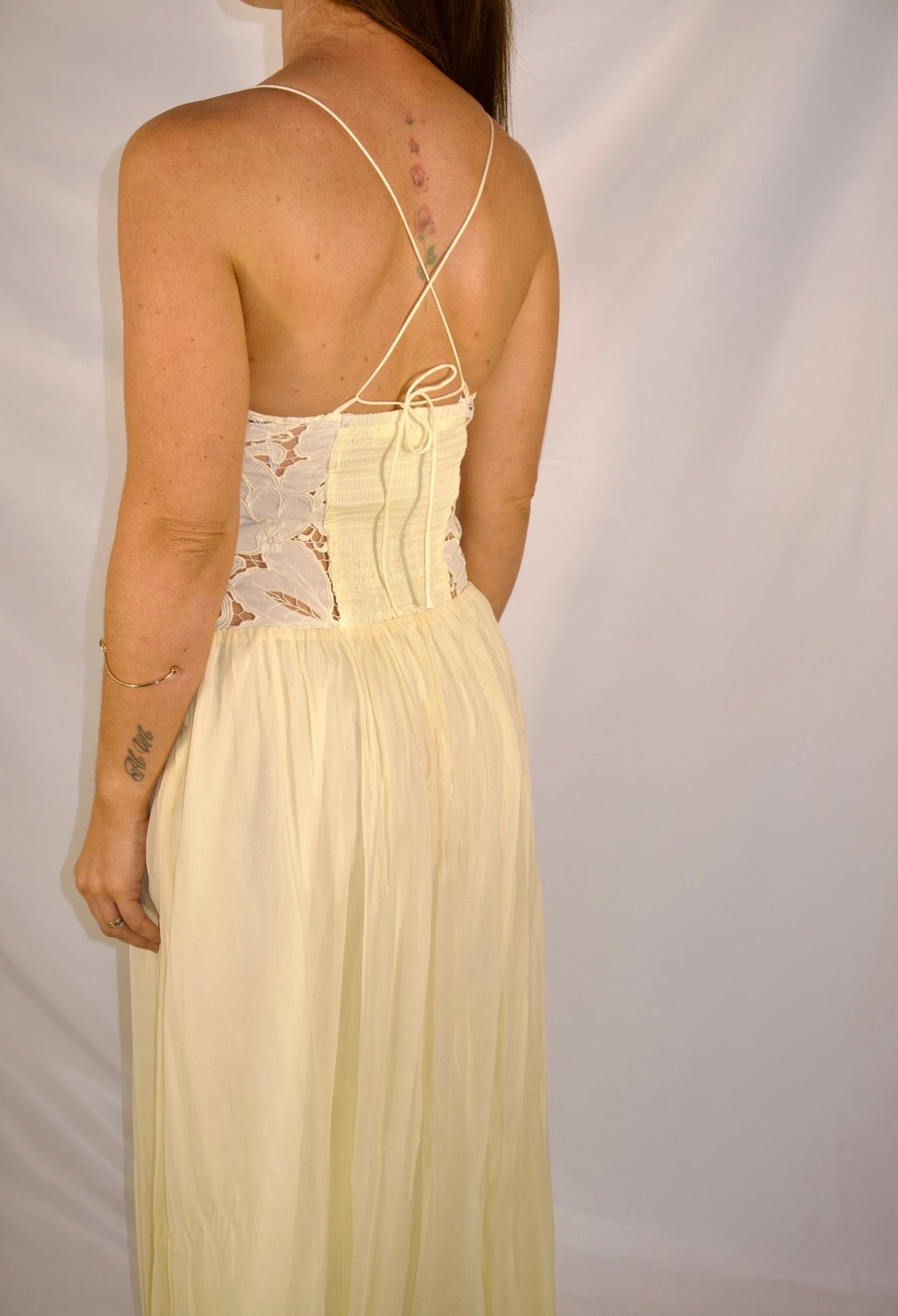 A woman shown from the back wearing a pale yellow maxi dress with a lace corset-style back bodice and thin tie-back straps, photographed against a plain light backdrop.