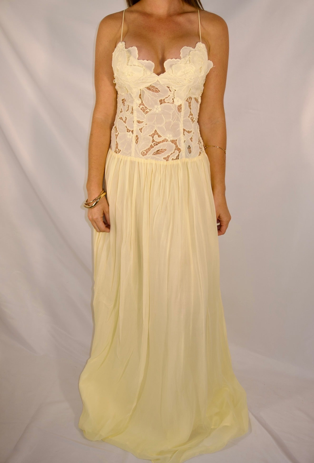 A woman modeling a long cream or pale yellow maxi dress with a floral lace bodice, thin straps, and a flowing chiffon skirt, photographed against a white draped backdrop.