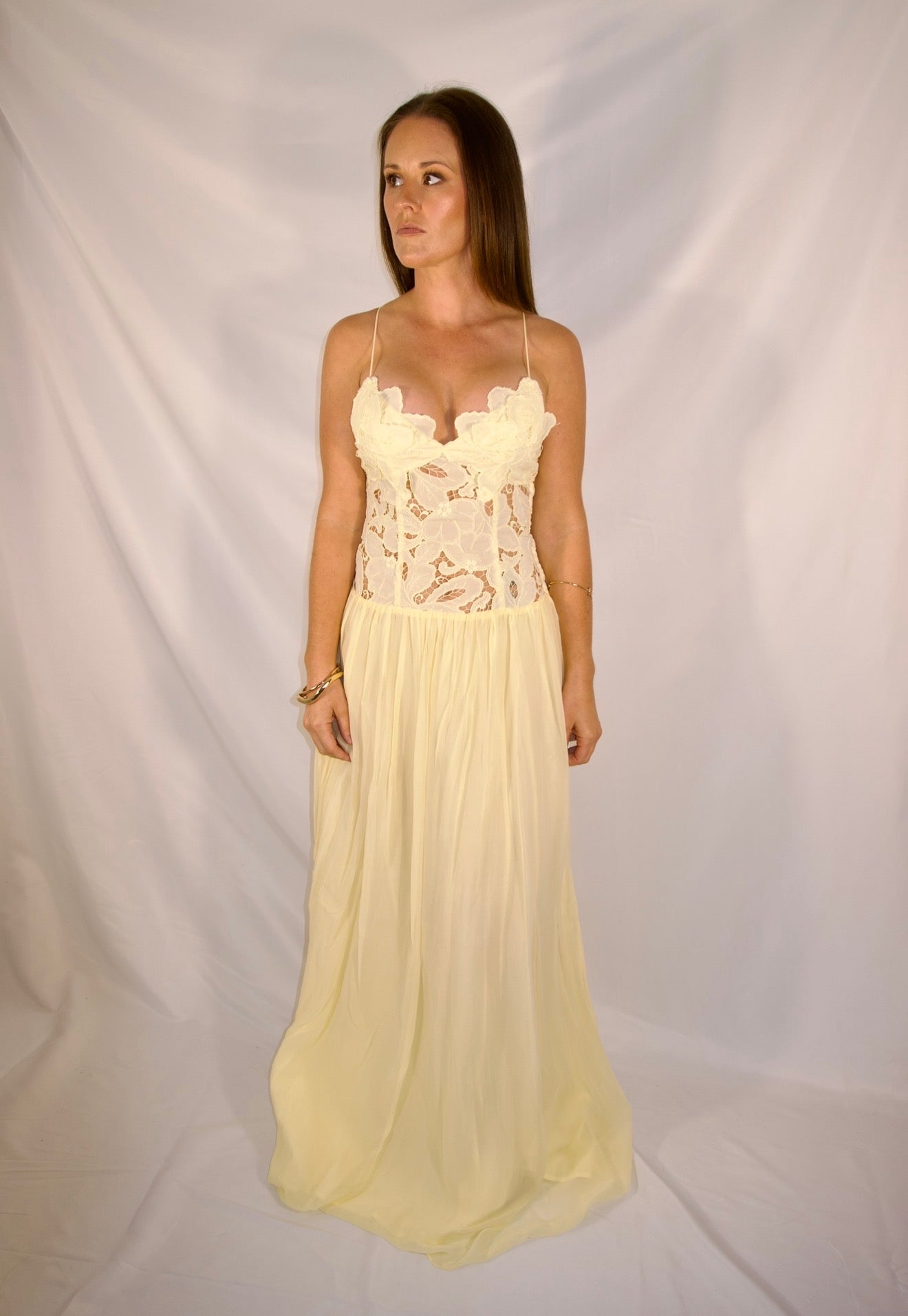 A woman modeling a long cream or pale yellow lace and mesh maxi dress with thin straps and a flowing skirt, standing against a white draped fabric backdrop.