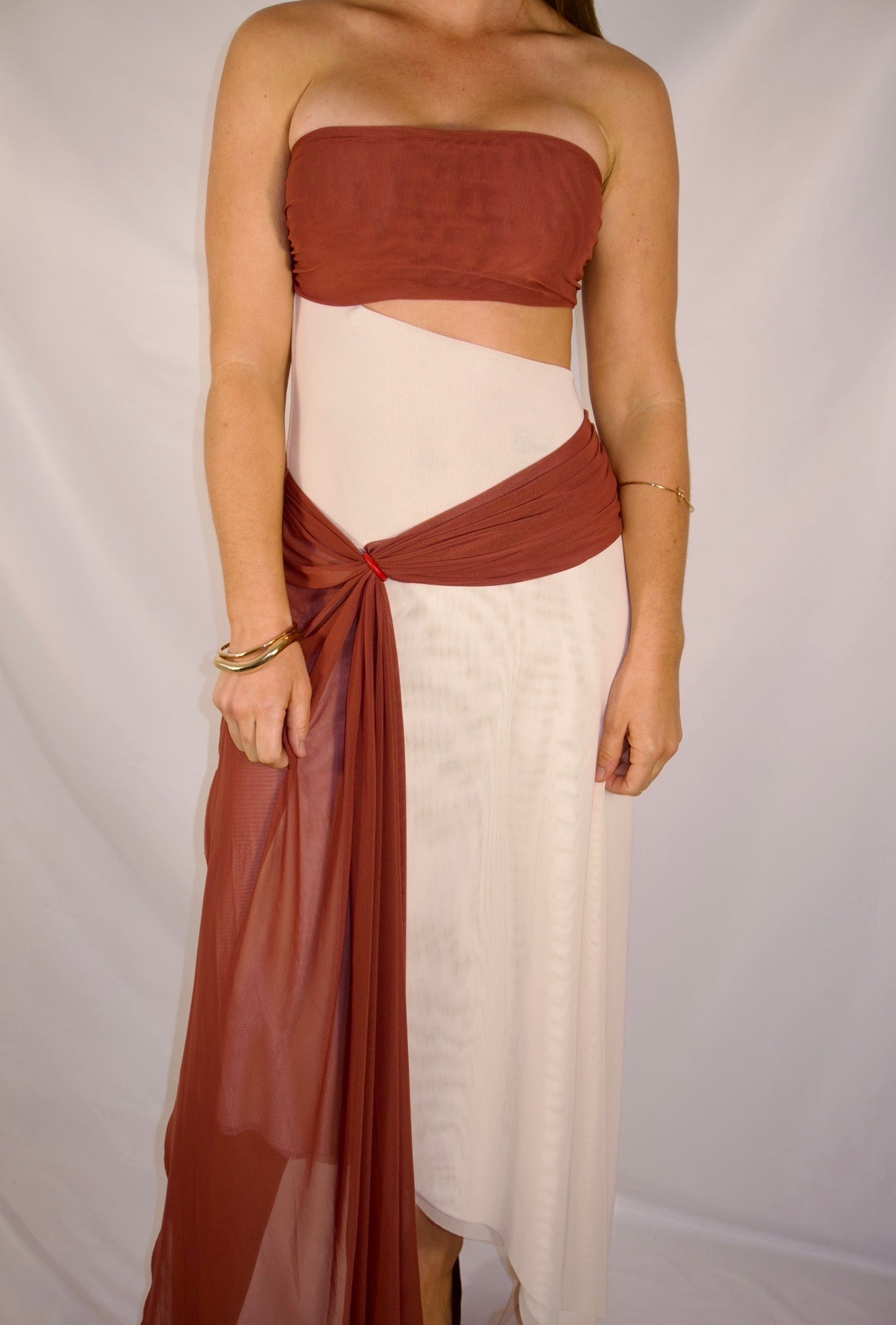 A woman modeling a two-piece draped dress with a terracotta rust top and matching draped skirt panels over an off-white underlayer, set against a white studio backdrop.