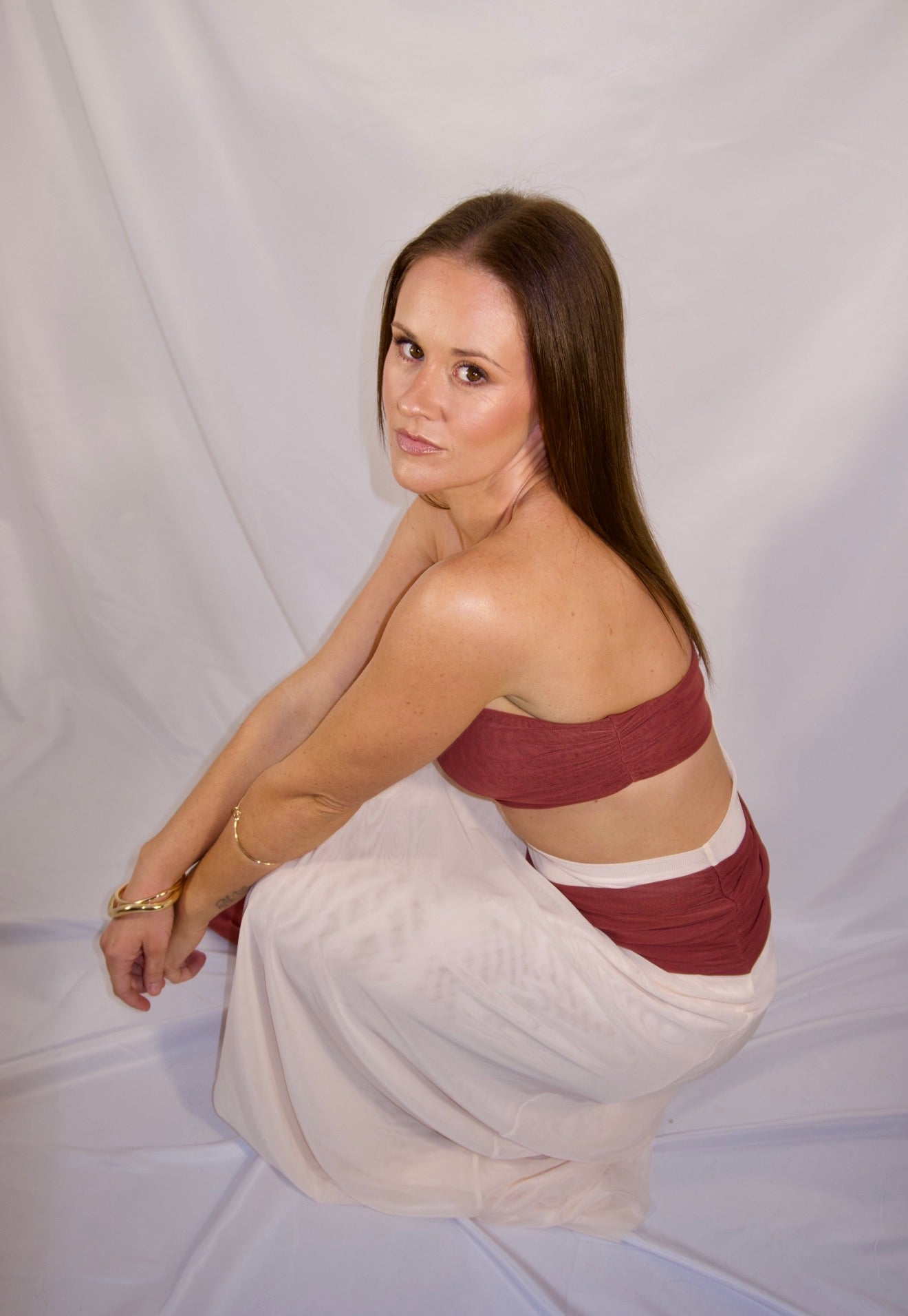 A woman wearing a two-piece set consisting of a maroon bandeau top and a matching maroon-and-white skirt, posing against a white fabric backdrop.
