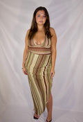 A woman modeling a sleeveless, wrap-style maxi dress with vertical brown, beige, and olive green stripes, standing against a white fabric backdrop.