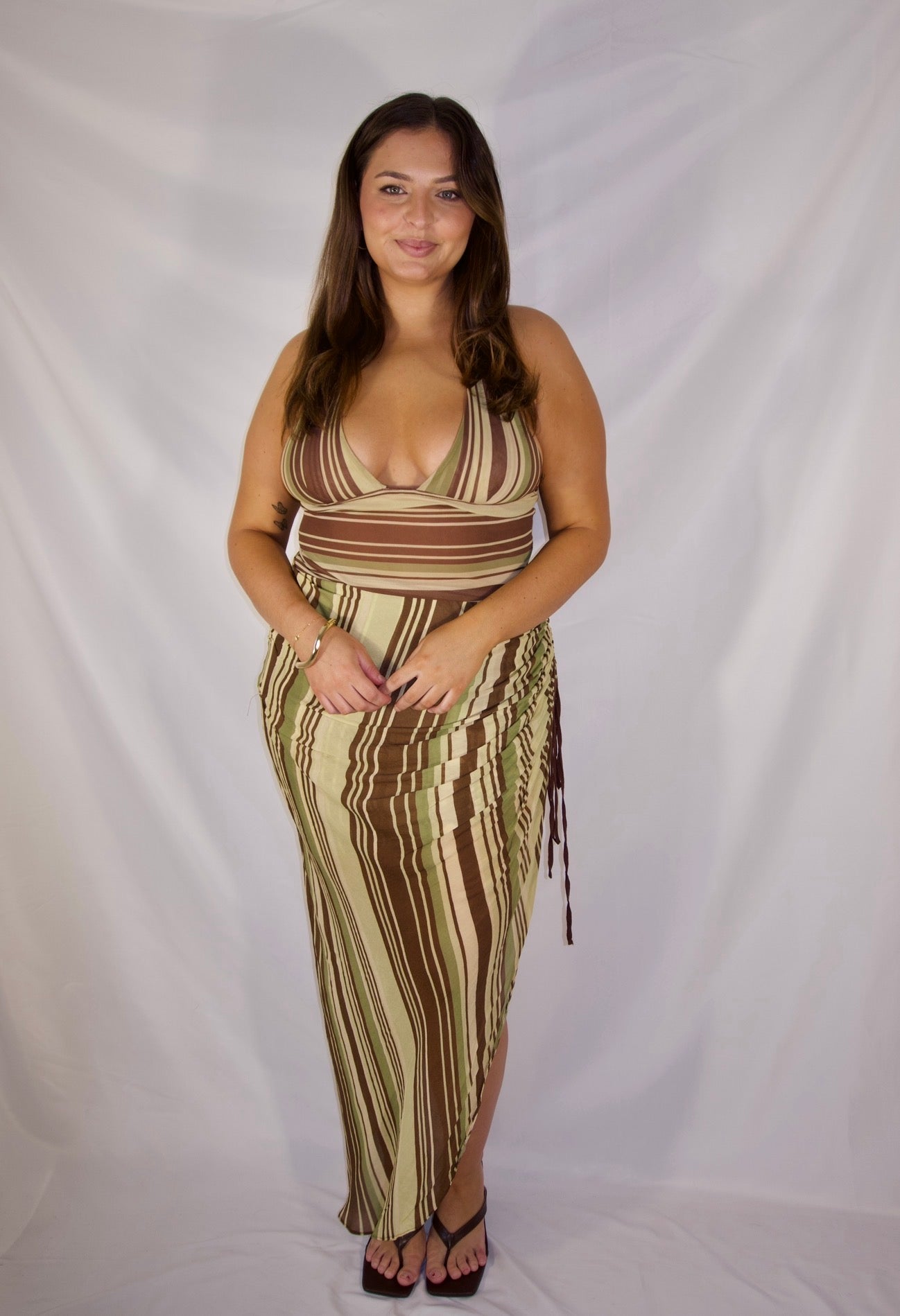 A woman modeling a sleeveless, wrap-style maxi dress with vertical brown and beige stripes, standing against a white fabric backdrop.
