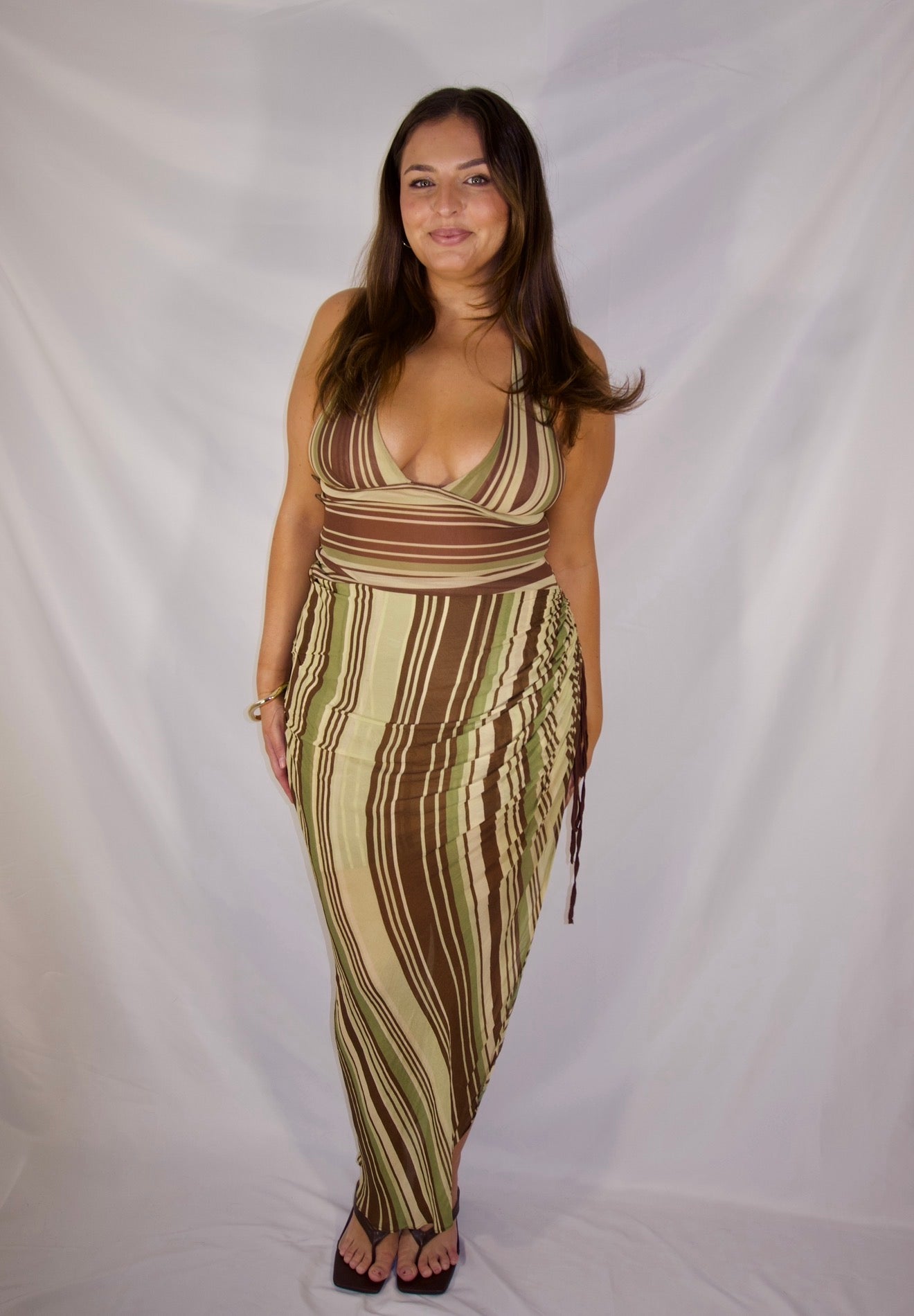A woman modeling a sleeveless wrap maxi dress with vertical brown, beige, and olive green stripes, standing against a white fabric backdrop.