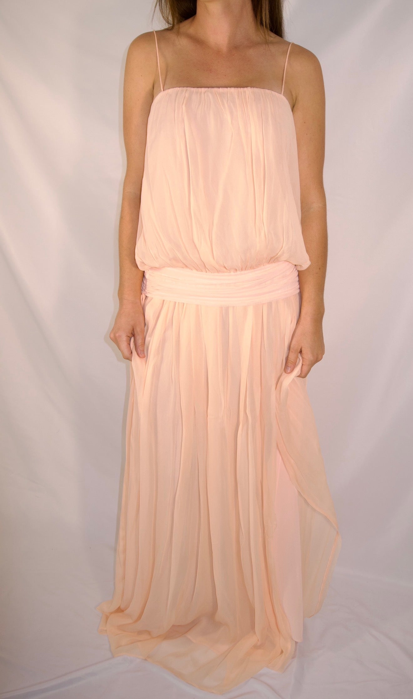A woman wearing a pale peach chiffon maxi dress with spaghetti straps and a matching fabric sash at the waist, standing against a white draped backdrop.