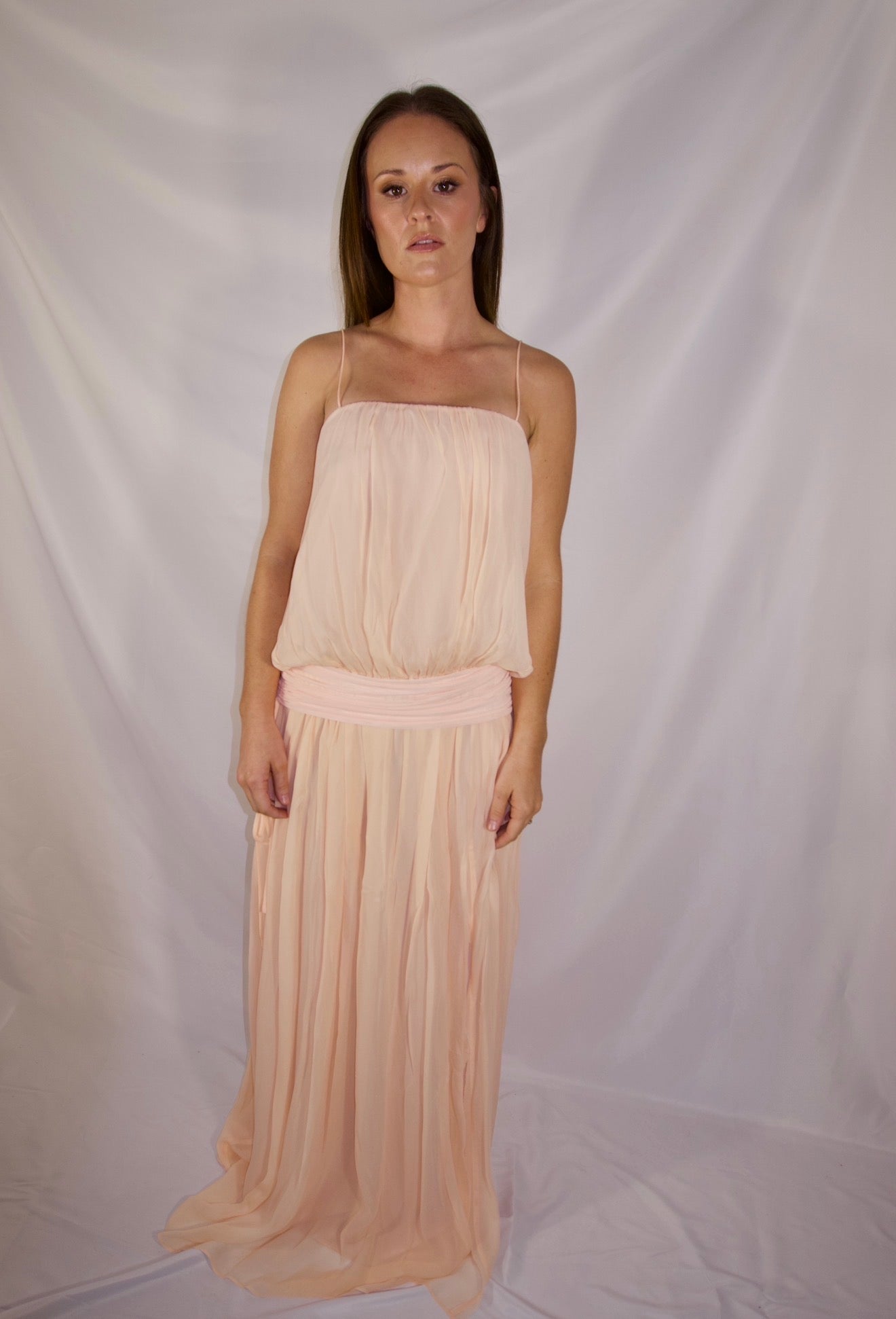 A woman modeling a sleeveless, floor-length peach chiffon dress with spaghetti straps and a matching fabric sash at the waist, set against a white draped fabric backdrop.