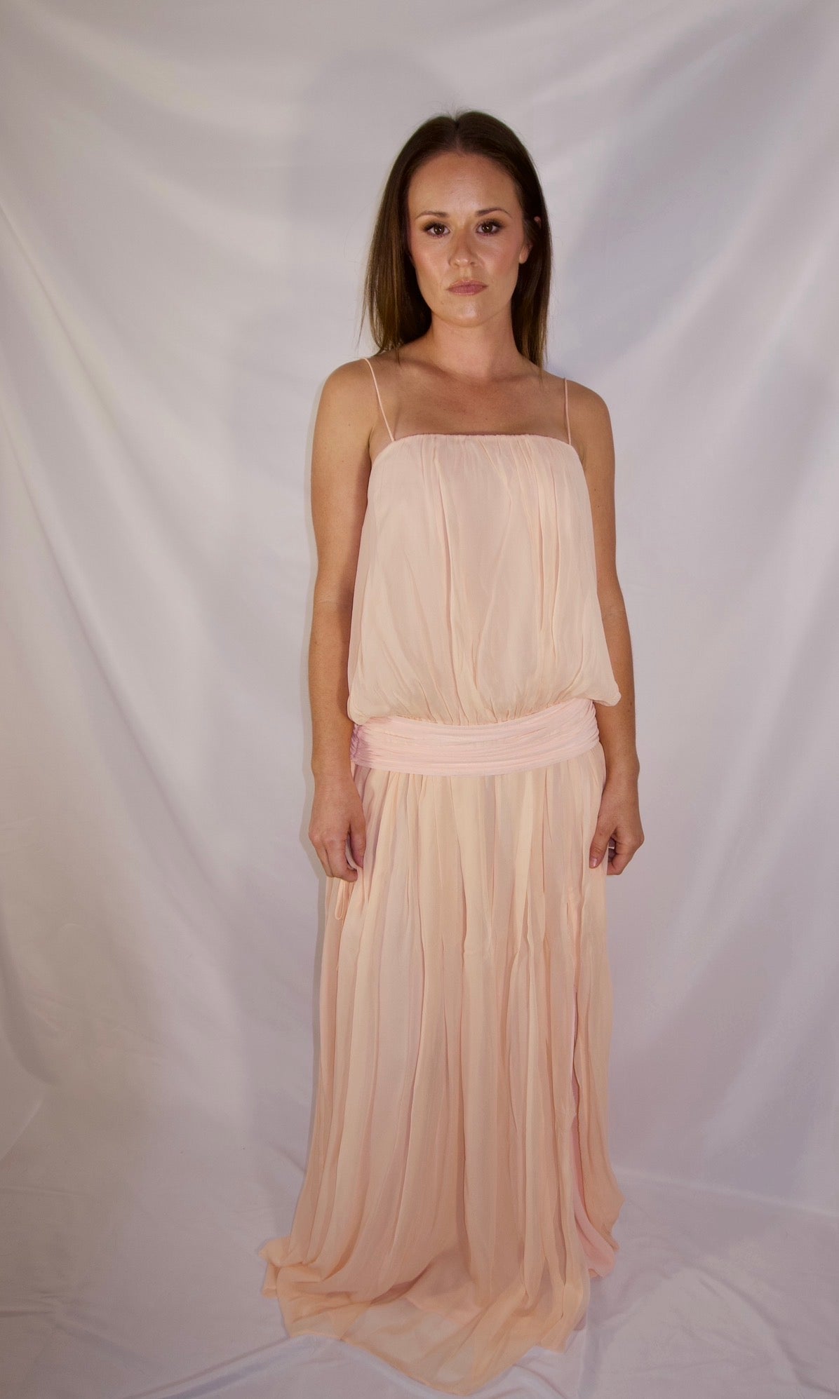 A woman modeling a light peach chiffon maxi dress with spaghetti straps and a matching fabric sash at the waist, standing against a white draped studio backdrop.