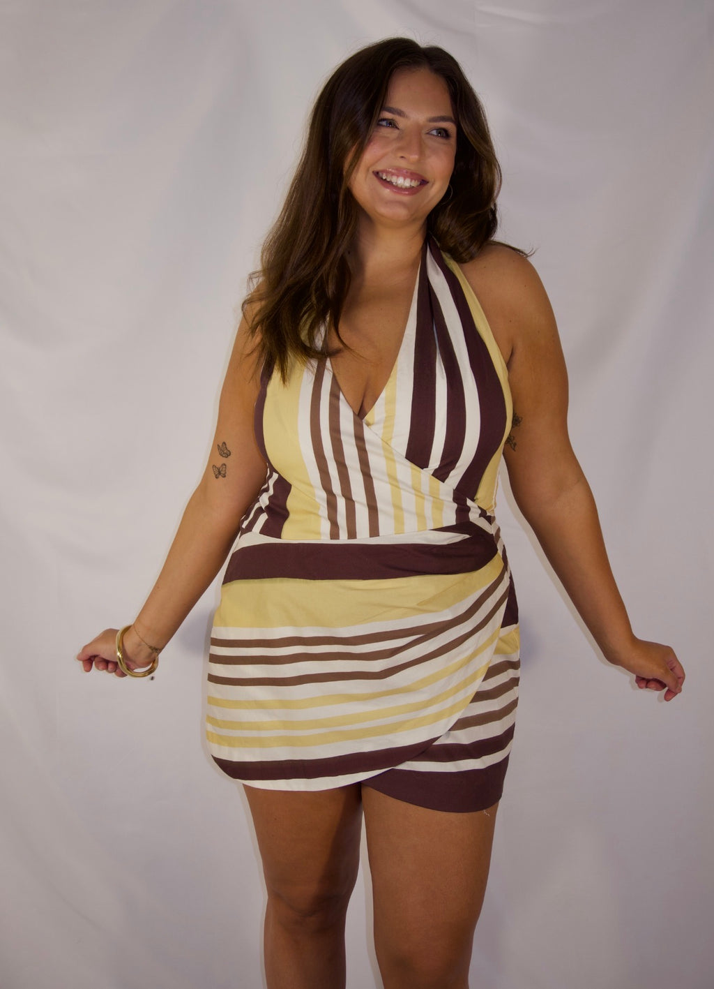 A woman modeling a halter-neck mini dress with a retro striped pattern in yellow, brown, and white, posing against a white studio backdrop.