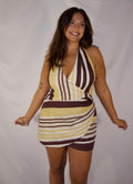 A woman modeling a halter-neck mini dress with a retro striped pattern in yellow, brown, and white, posing against a white studio backdrop.