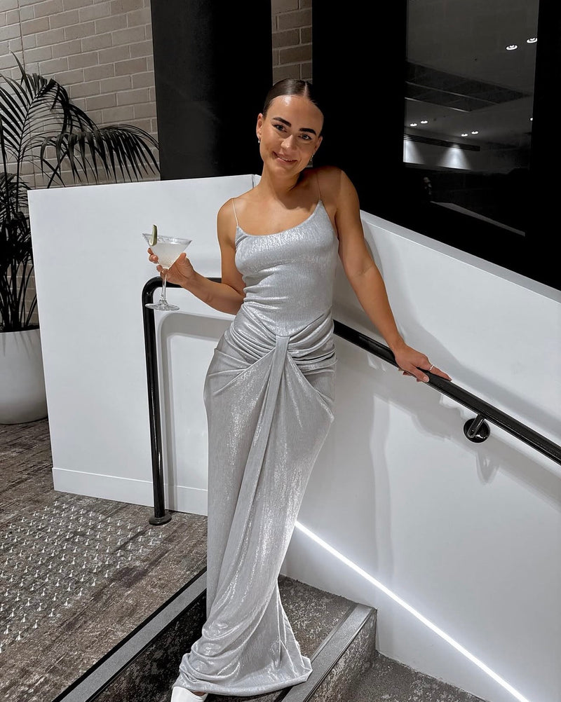 A woman wearing a shimmering silver spaghetti-strap evening gown with a draped twist detail at the waist, posing on a modern white staircase while holding a martini glass. - @paytonmortimer_