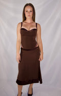 A woman modeling a dark brown sleeveless slip dress with a sweetheart neckline, sheer mesh cups, side cutouts, and a matching skirt, standing against a white draped studio backdrop.