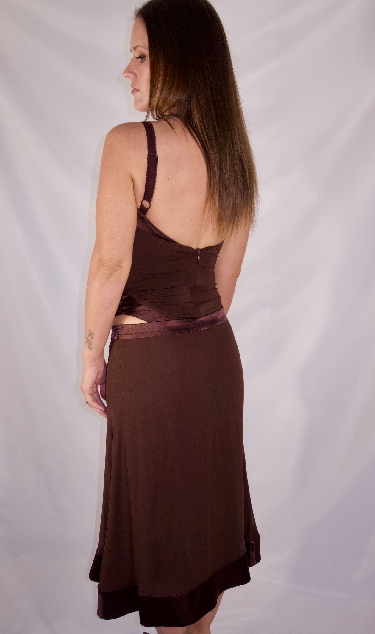 A woman modeling a dark brown sleeveless dress from the back, standing against a white fabric backdrop.