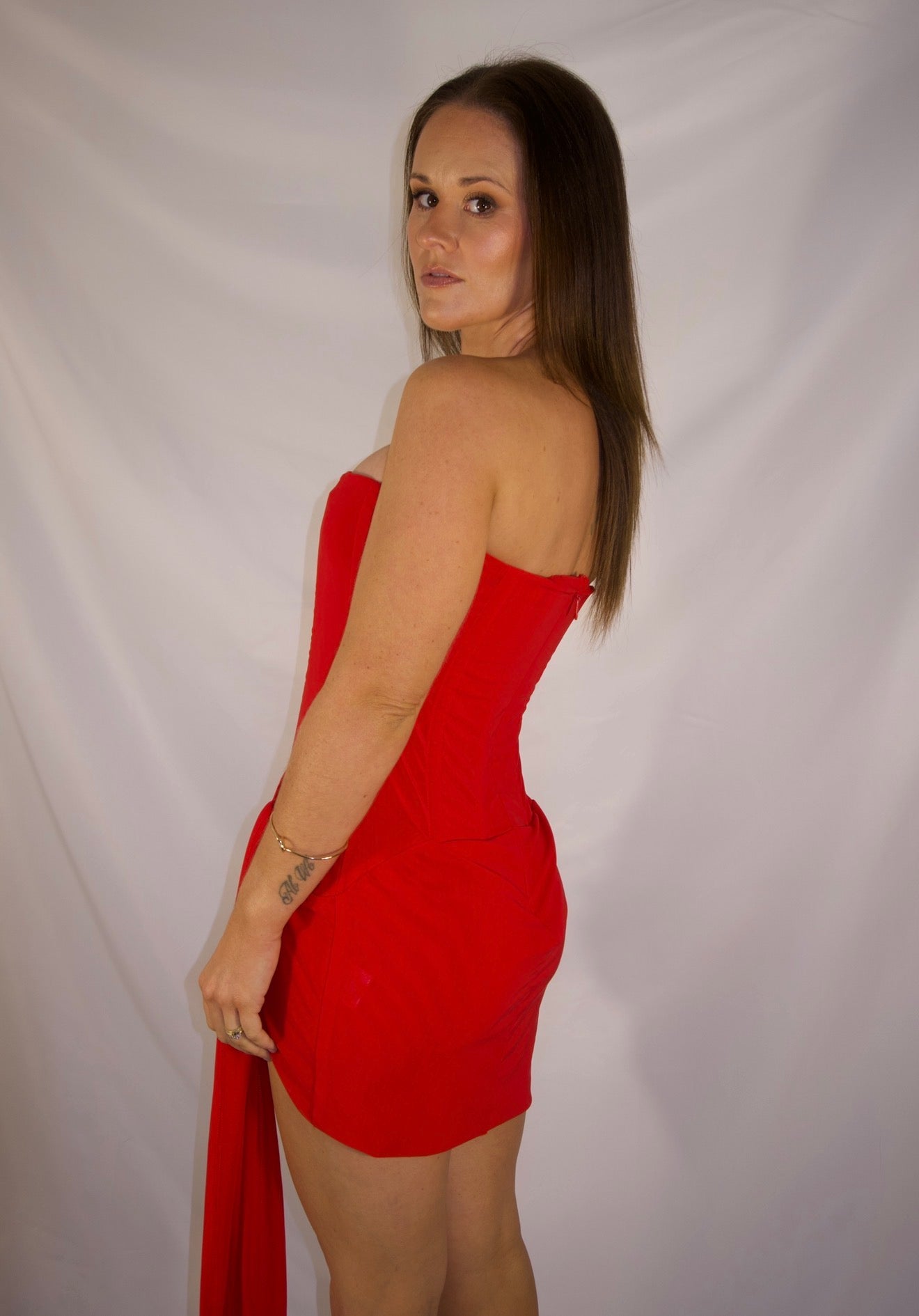 A woman modeling a strapless red mini dress with a long flowing train, photographed against a white fabric backdrop.
