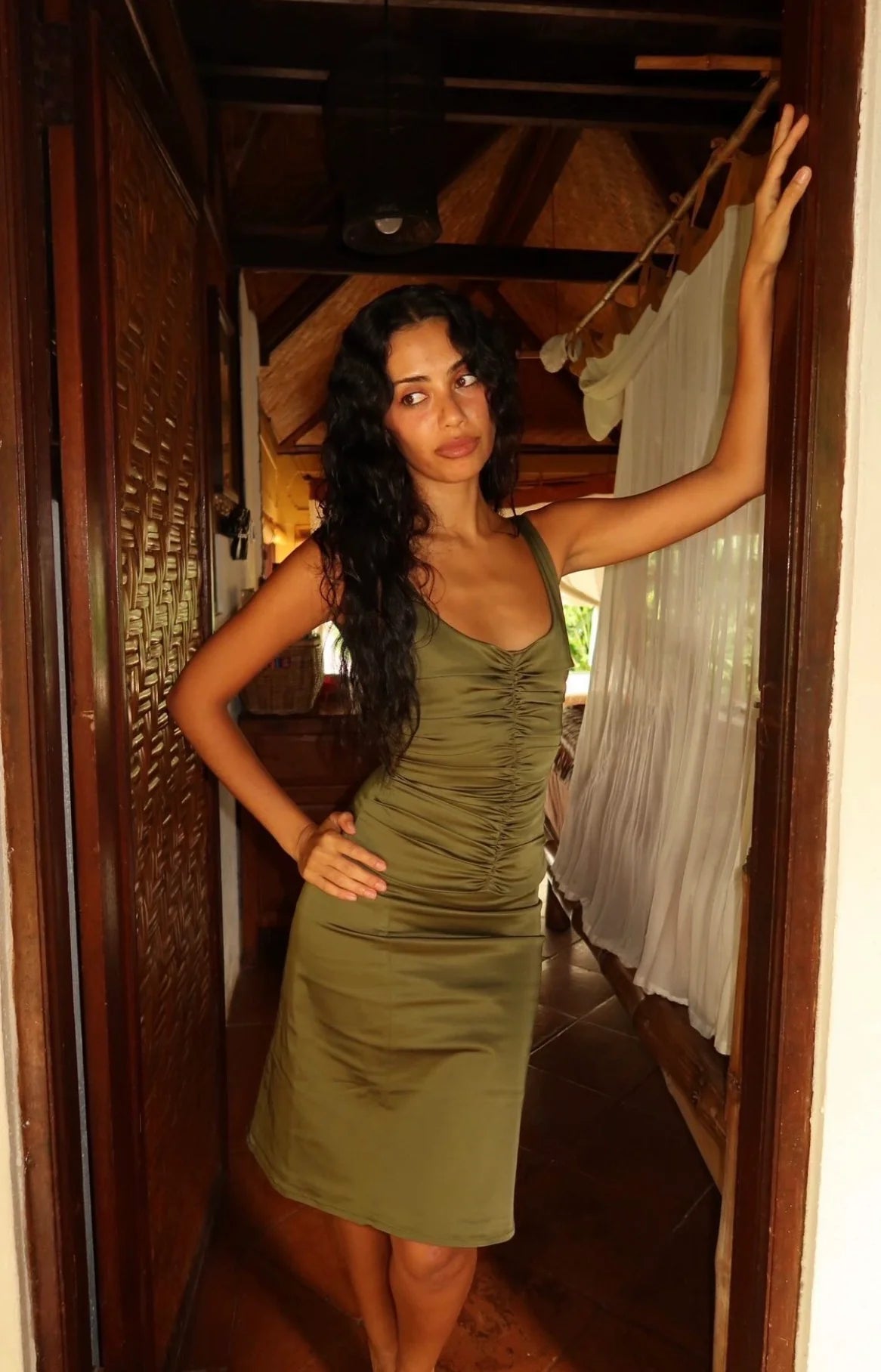A woman with long dark hair wearing a sleeveless olive green ruched mini dress, posing in a doorway with wooden walls and white curtains in the background.