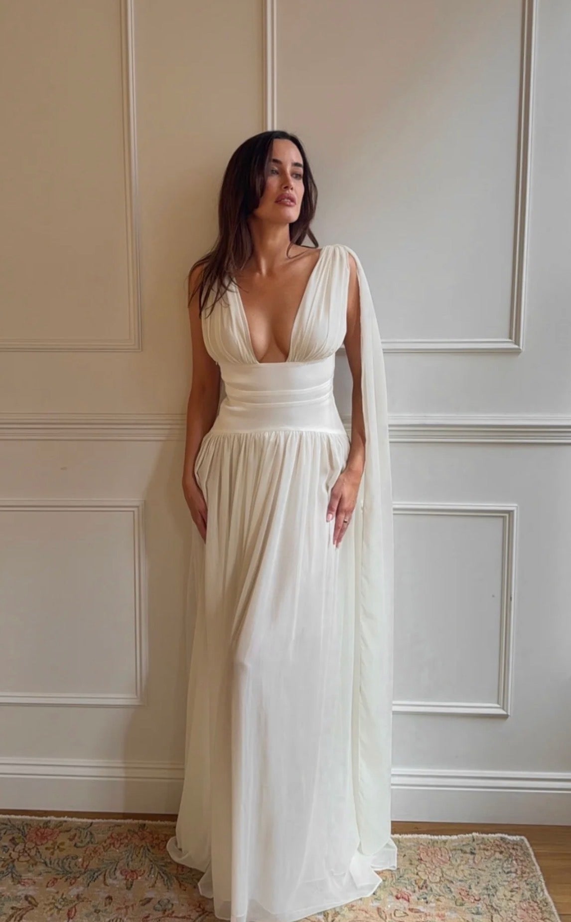 A woman wearing a floor-length ivory wedding dress with a deep plunging neckline, a fitted waist, and a flowing sheer cape-like overlay, standing indoors against a cream paneled wall.