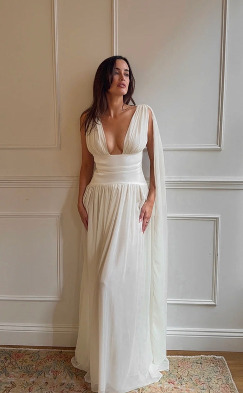 A woman wearing a floor-length ivory wedding dress with a deep plunging neckline, a fitted waist, and a flowing sheer cape-like overlay, standing indoors against a cream paneled wall.