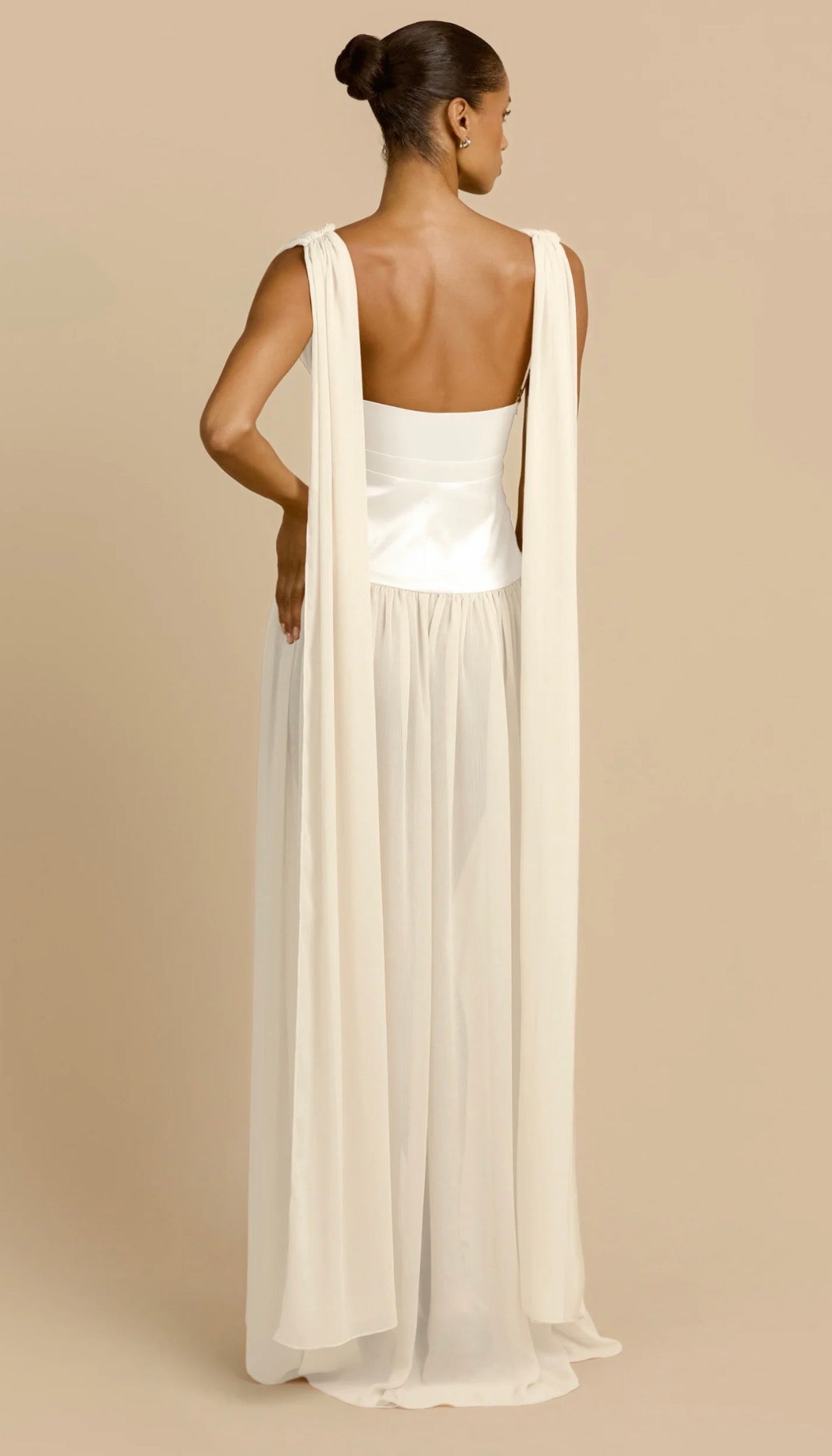 A woman shown from the back wearing a floor-length white dress with a satin-like bodice, wide shoulder straps, and a flowing sheer overlay, photographed against a plain warm beige studio background.