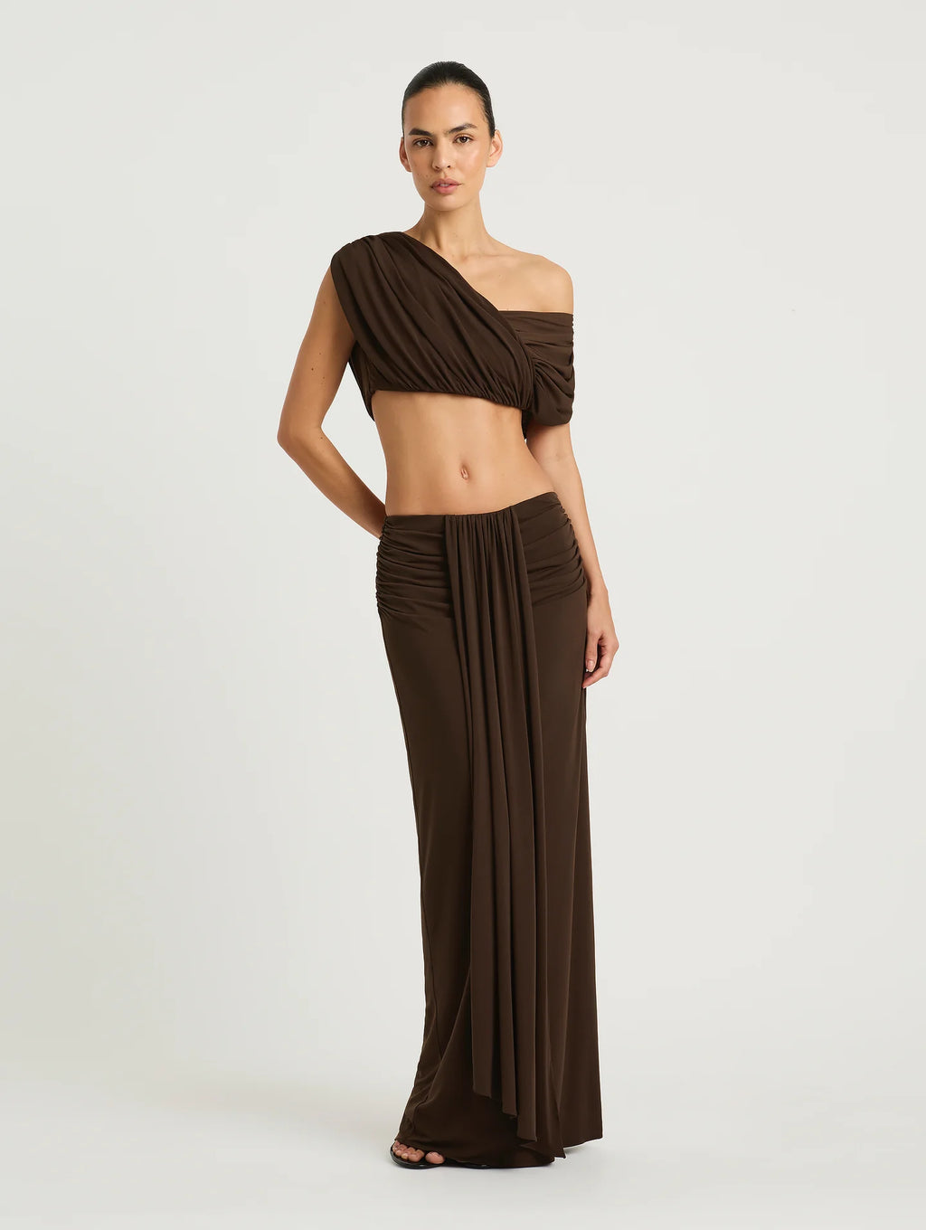 A model wearing a dark brown two-piece set consisting of a draped off-the-shoulder crop top and matching pleated maxi skirt, photographed against a light gray studio background.