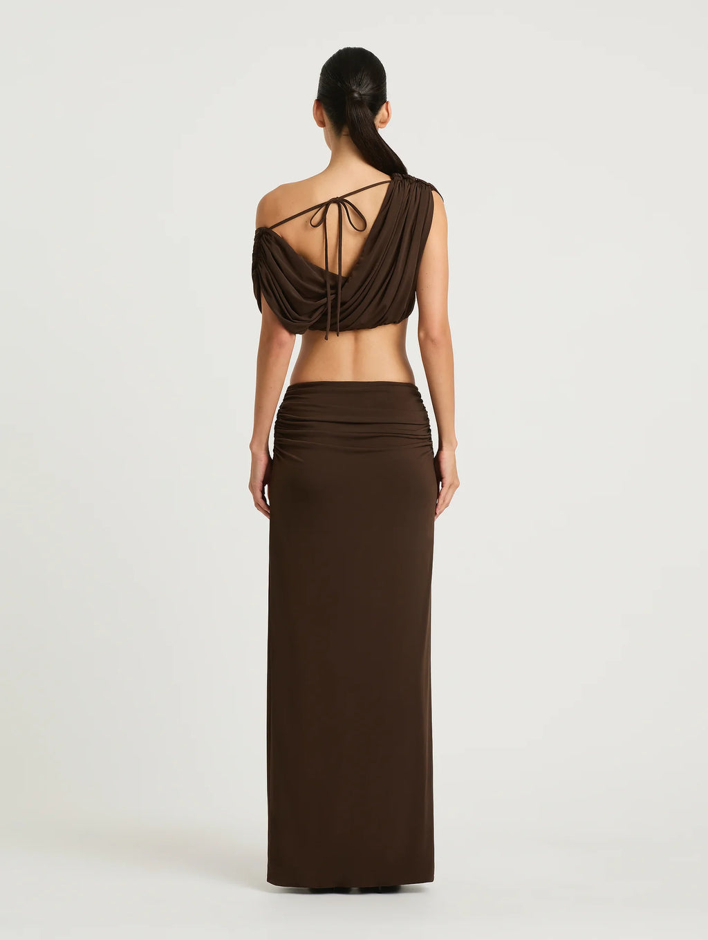 A woman shown from the back wearing a dark brown two-piece outfit consisting of a one-shoulder draped crop top with tie details and a matching long skirt, against a light gray studio background.