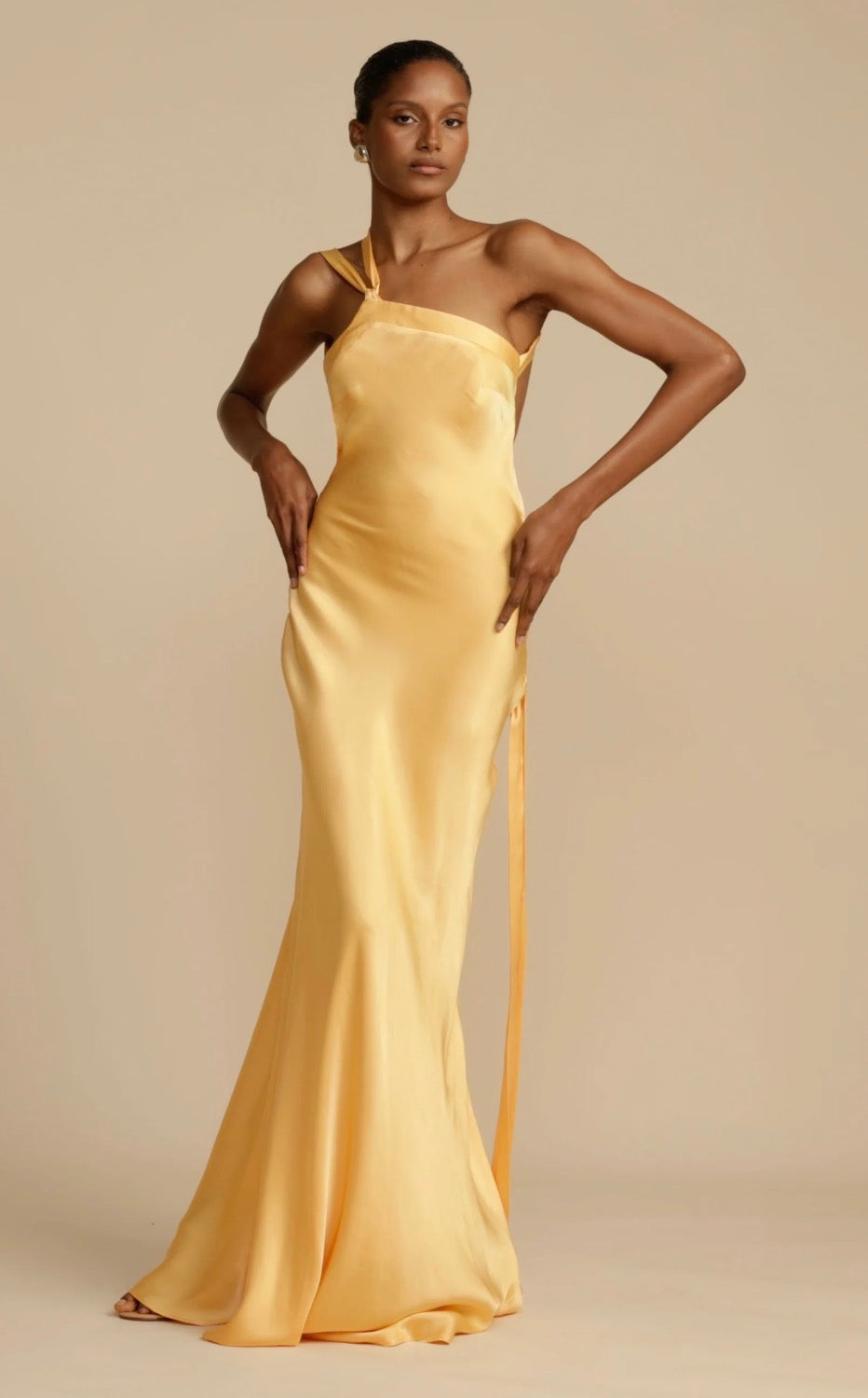 A woman modeling a pale yellow satin one-shoulder evening gown with a side tie, set against a solid beige studio background.