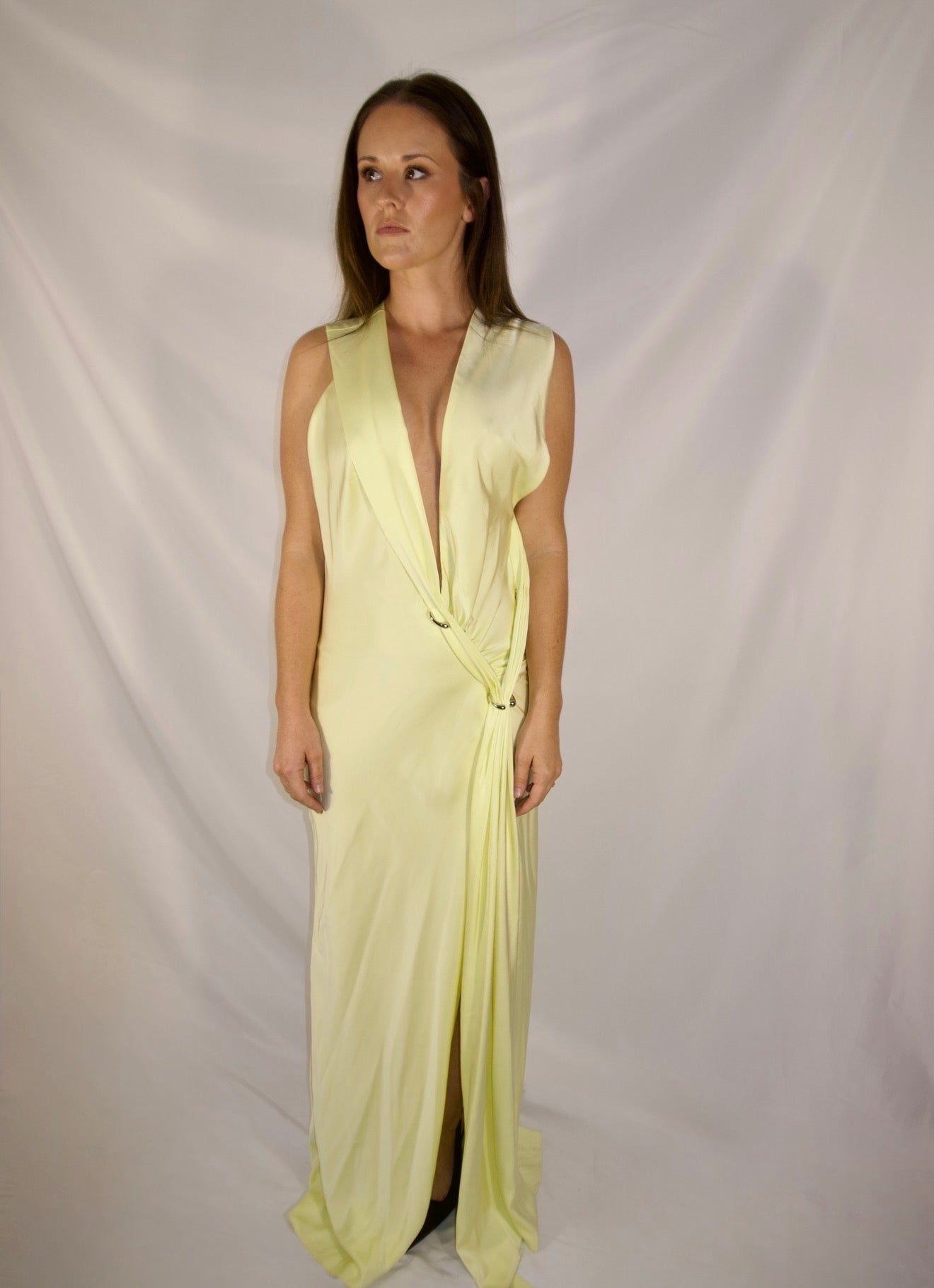 A woman modeling a pale yellow sleeveless wrap maxi dress with a deep plunging V-neckline and a twisted waist detail, set against a draped white fabric backdrop.