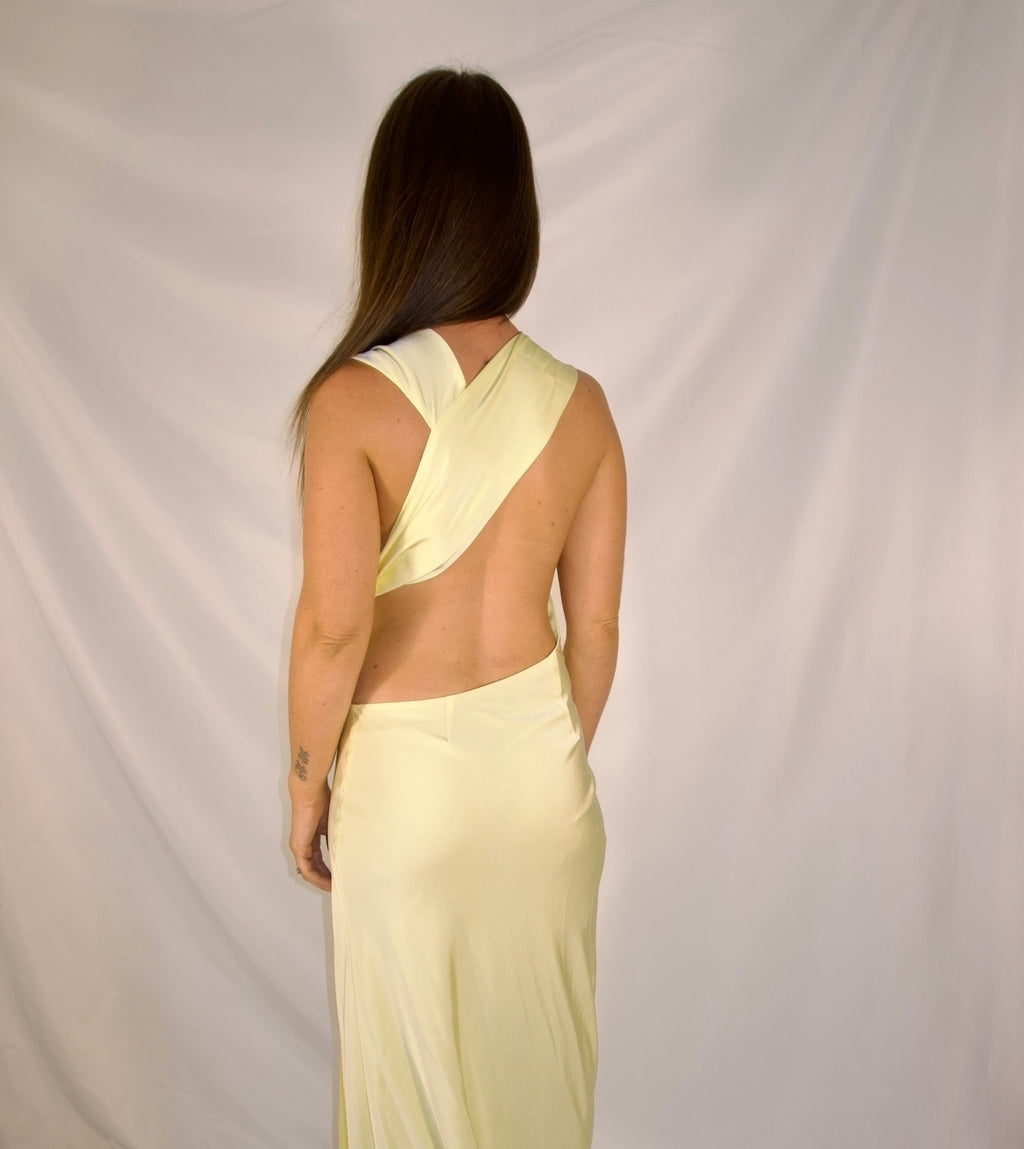 A woman viewed from the back wearing a pale yellow satin dress with a draped crossover back design and a low open back, set against a draped white fabric backdrop.