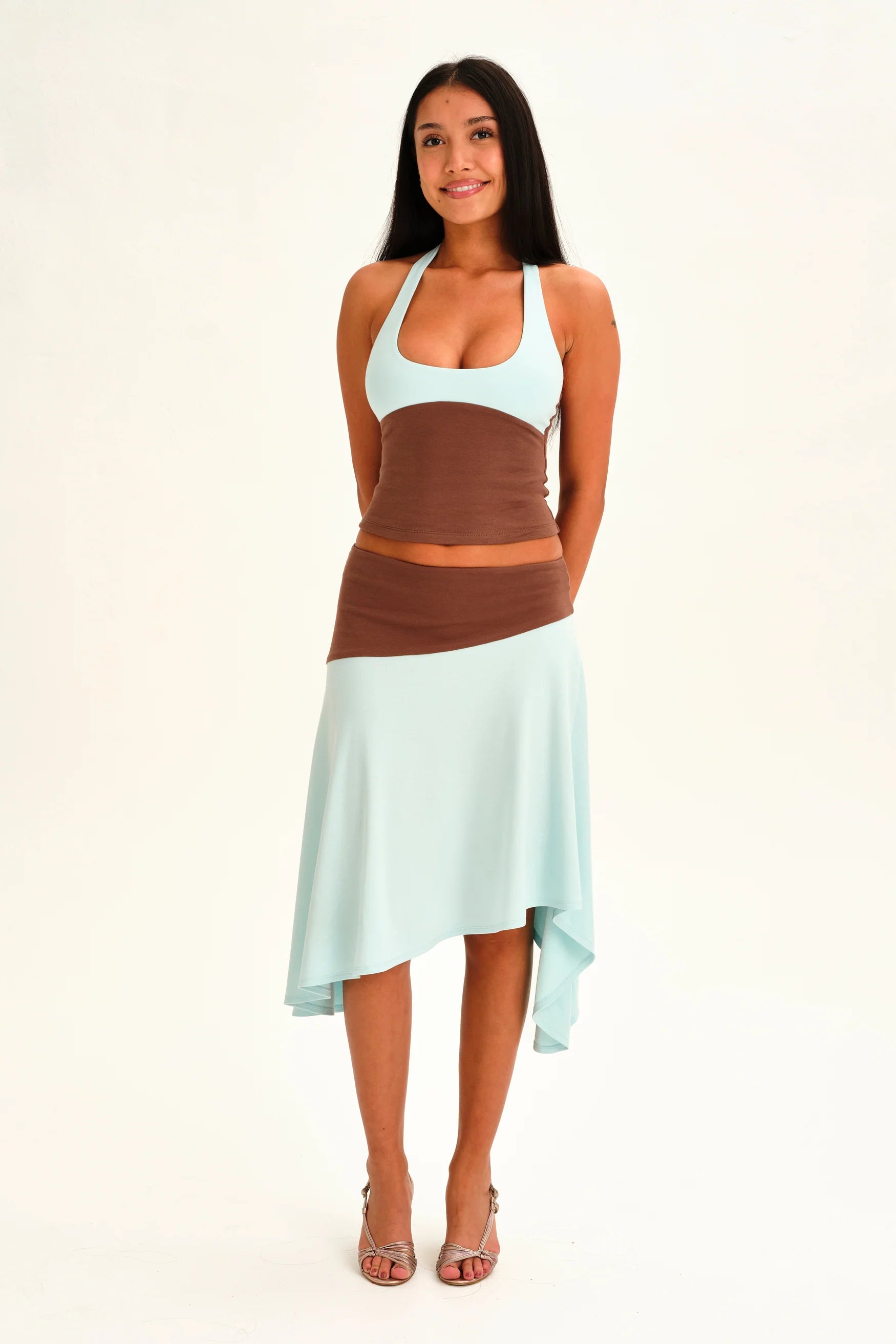 A woman modeling a color-blocked knit two-piece set featuring a mint green halter top and matching high-low skirt with brown color-blocked panels, set against a plain white studio background.