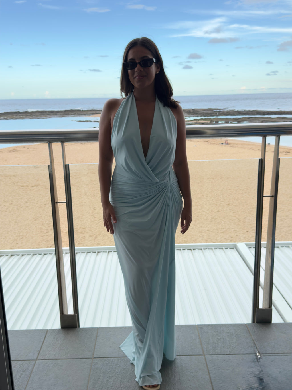 Woman in a light blue dress standing on a balcony with a beach view. - @belle.govindaley