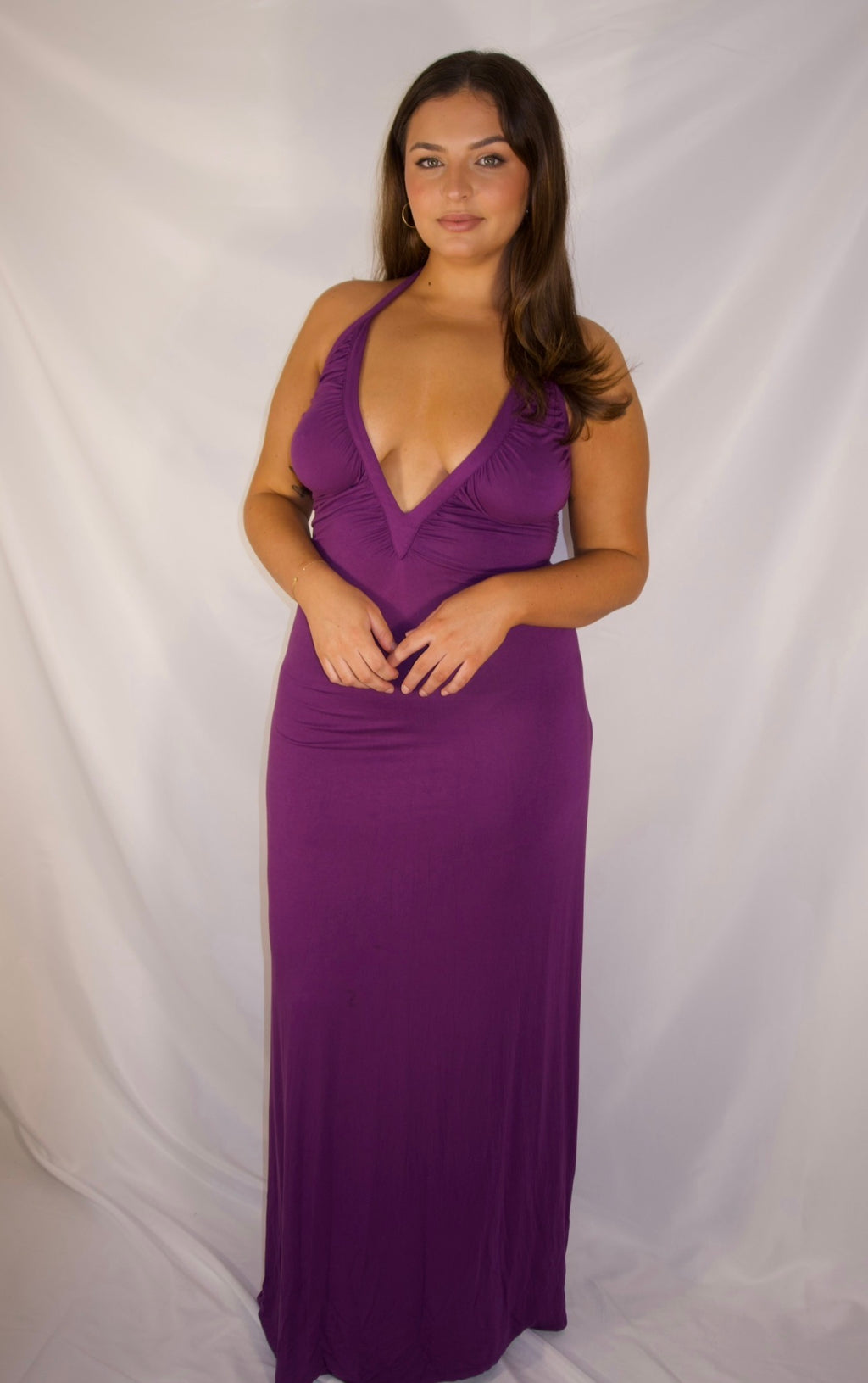 A woman modeling a sleeveless, deep V-neck, floor-length purple maxi dress against a white fabric backdrop.