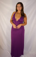 A woman modeling a sleeveless, deep V-neck, floor-length purple maxi dress against a white fabric backdrop.