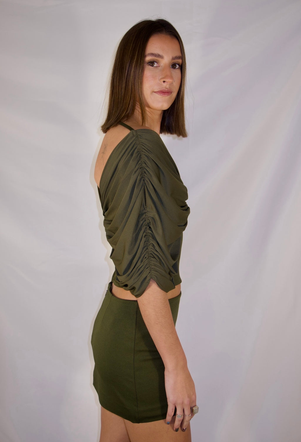 A woman wearing an olive green ruched crop top and matching mini skirt, photographed against a white studio backdrop. - @_ellahbates