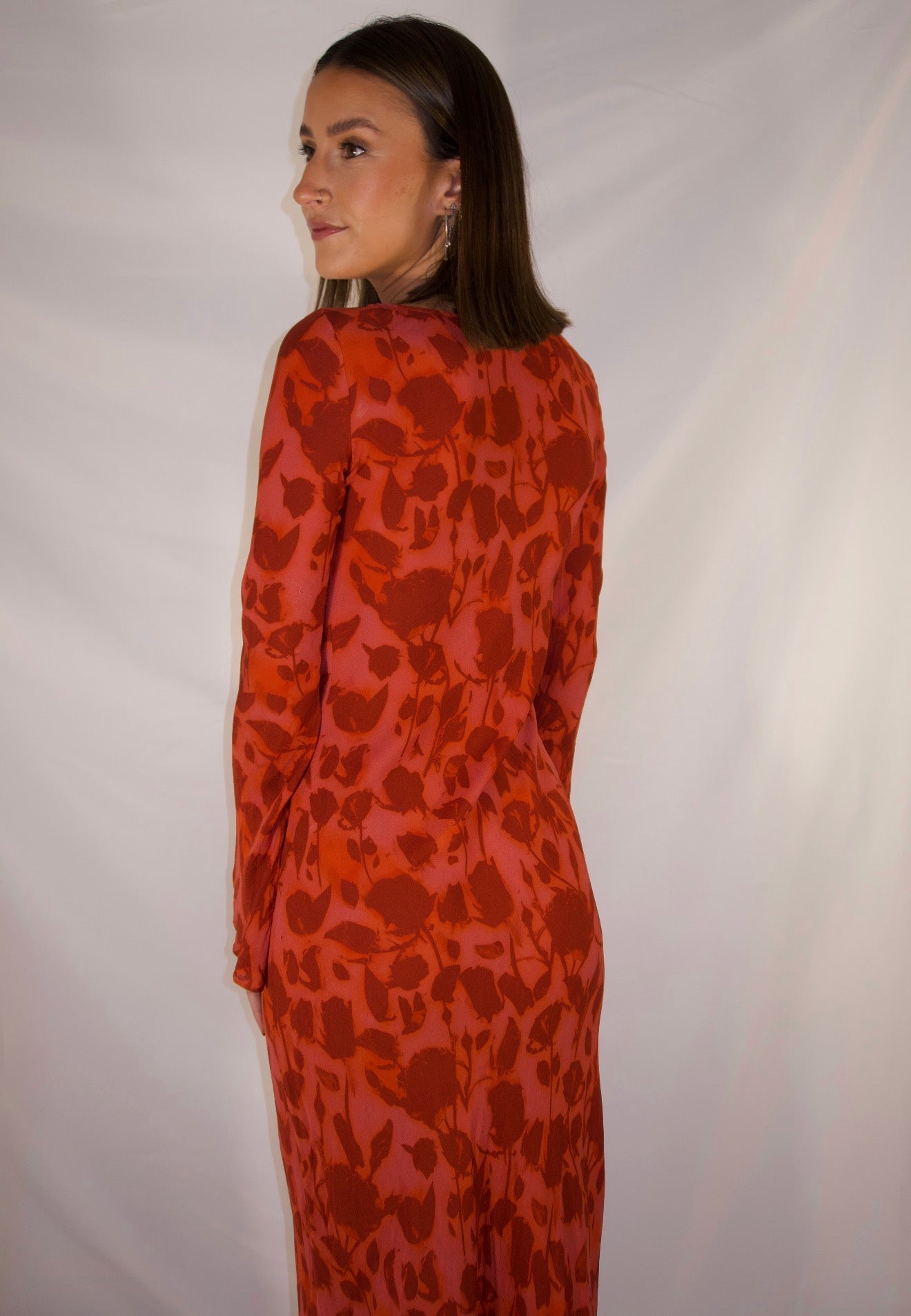 A woman wearing a long-sleeve red dress with a dark red abstract floral pattern, photographed from the back against a white studio backdrop.