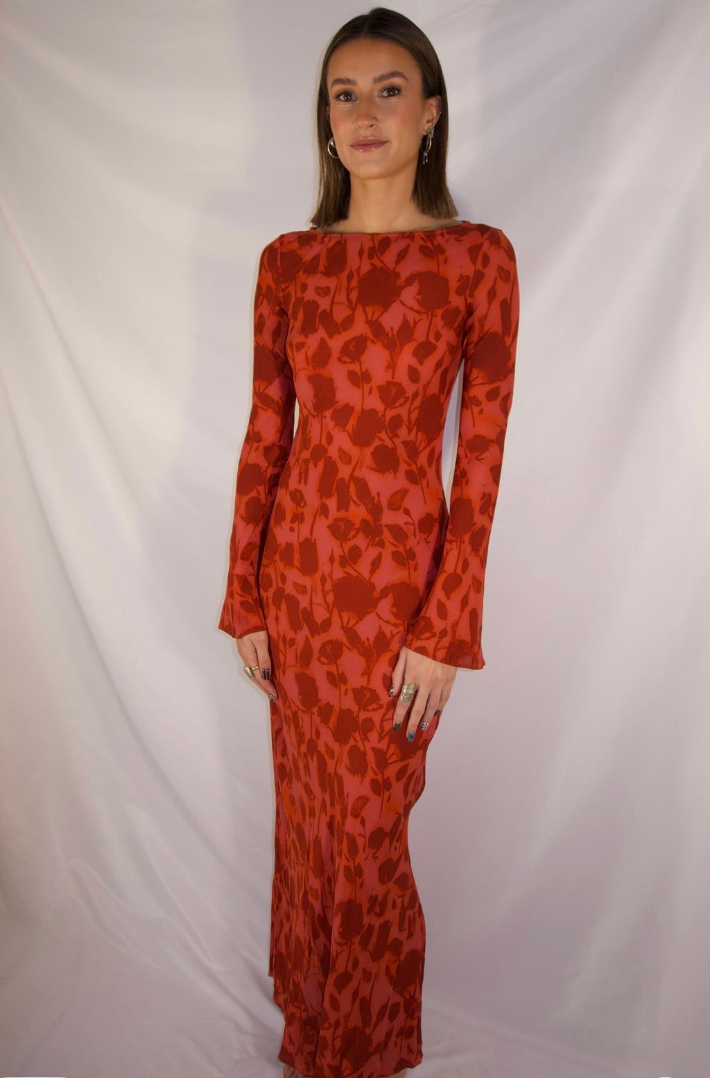 A woman modeling a long-sleeved, floor-length red dress with a dark red abstract floral pattern, standing against a white draped fabric backdrop.