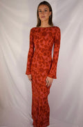 A woman modeling a long-sleeved, floor-length red dress with a dark red abstract floral pattern, standing against a white draped fabric backdrop.